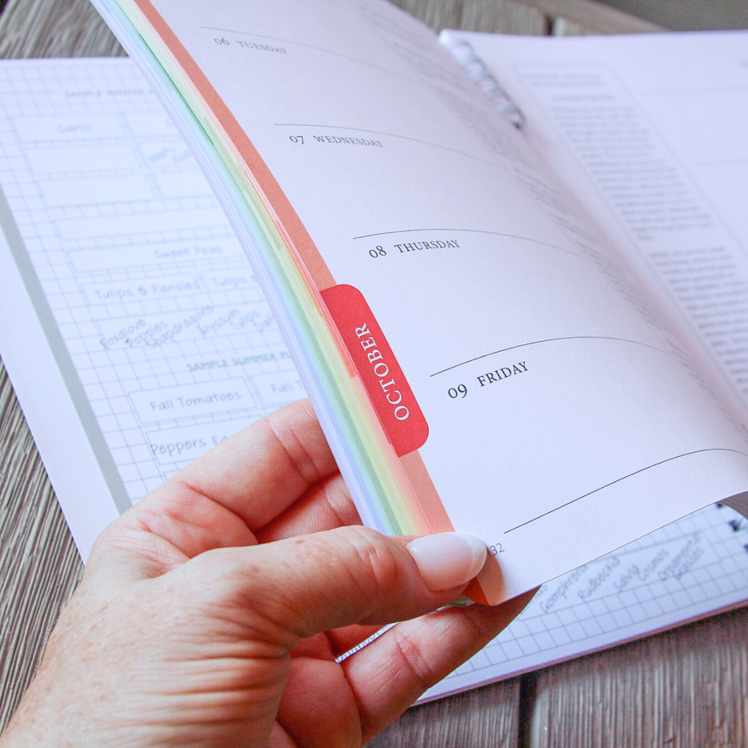 Hand holding the colorful North Texas Garden Planner with a red 'October' tab, open to a page on a wooden surface.